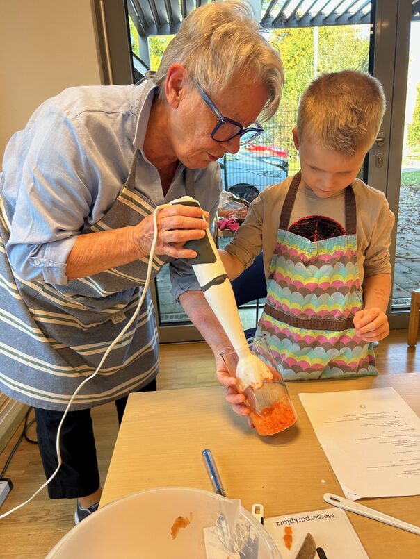 Eine ältere Frau und ein Kind stehen an einem Tisch. Die Frau hat einen Pürierstab und einen Becher mit zerkleinerten Möhren in der Hand