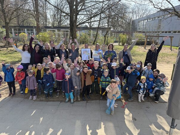 Eine Gruppe von Kita- Kindern und Erwachsenen stehen auf dem Spielplatz und halten die Hände in die Luft. In der Mitte halten zwei Frauen einen Bilderrahmen mit dem KITA Vital Zertifikat in den Händen.