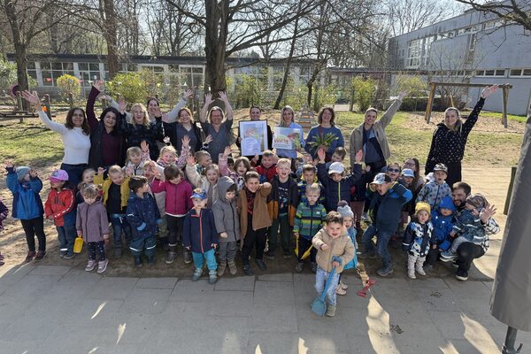 Eine Gruppe von Kita- Kindern und Erwachsenen stehen auf dem Spielplatz und halten die Hände in die Luft. In der Mitte halten zwei Frauen einen Bilderrahmen mit dem KITA Vital Zertifikat in den Händen.