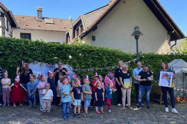 Eine Gruppe von Kindern und Erwachsenen steht vor einer Hecke . Rechts im Bild hält eine Frau einen Bilderrahmen mit einem Zertifikat in den Händen.