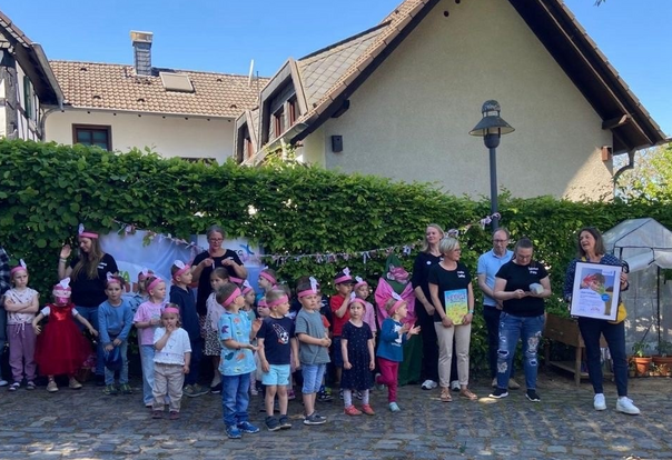 Eine Gruppe von Kindern und Erwachsenen steht vor einer Hecke . Rechts im Bild hält eine Frau einen Bilderrahmen mit einem Zertifikat in den Händen.