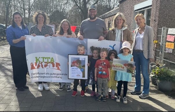Erwachsene halten ein Banner mit der Aufschrift KITA Vital in die Höhe und einige Kinder stehen davor
