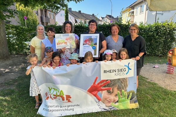 Kinder und Erwachsene stehen auf einer Wiese und halten ein Banner vor sich auf dem das Gesicht eines Jungen mit einer erhobenen grünen und roten Hand zu sehen ist