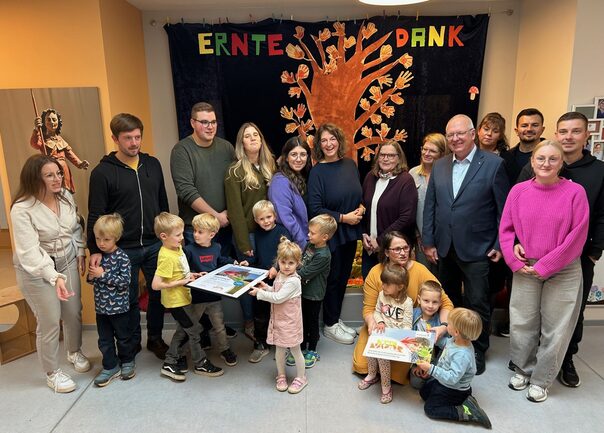 Eine Gruppe von Personen stehen vor einem Wandbehang auf dem Erntedank steht. Im Vordergrund stehen Kinder, die das KITA Vital Hausschild und das Zertifikat in den Händen halten.