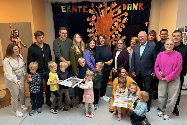 Eine Gruppe von Personen stehen vor einem Wandbehang auf dem Erntedank steht. Im Vordergrund stehen Kinder, die das KITA Vital Hausschild und das Zertifikat in den Händen halten.