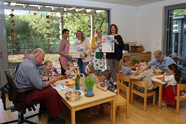 Im Vordergrund sitzen ein älterer Herr und eine Dame mit Kindern an jeweils einem Tisch , auf dem sich Küchenutensilien befinden. Im Hintergrund stehen vier Frauen, die ein Hausschild und ein Zertifikat in den Händen halten.