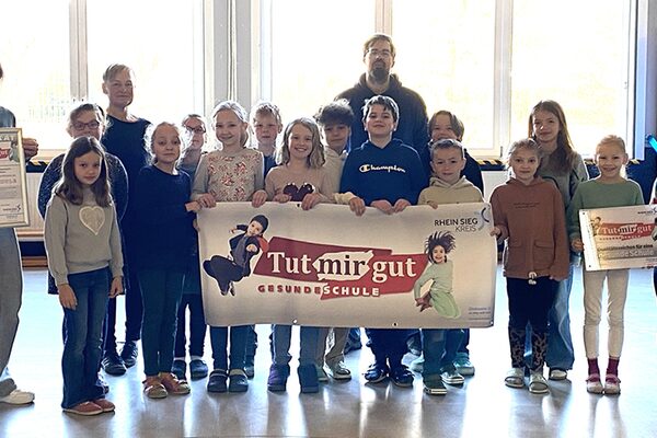 Eine Gruppe von Kindern steht in einer Turnhalle auf grauem Boden und hält ein Banner in der Hand. AUf diesem sind zwei hochspringende Kinder sichtbar. Am linken Bildrand steht eine Frau mit einem Bilderrahmen,und hinter der Kindergruppe steht eine weitere Frau und ein Mann.