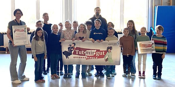 Eine Gruppe von Kindern steht in einer Turnhalle auf grauem Boden und hält ein Banner in der Hand. AUf diesem sind zwei hochspringende Kinder sichtbar. Am linken Bildrand steht eine Frau mit einem Bilderrahmen,und hinter der Kindergruppe steht eine weitere Frau und ein Mann.