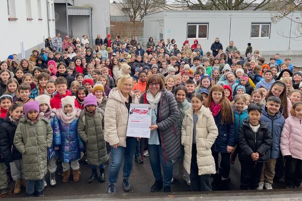Zwei Frauen stehen vor einer Traube von Kindern auf dem Schulhof einer Schule und halten gemeinsam einen Bilderrahmen mit einem Zertifikat in den Händen.