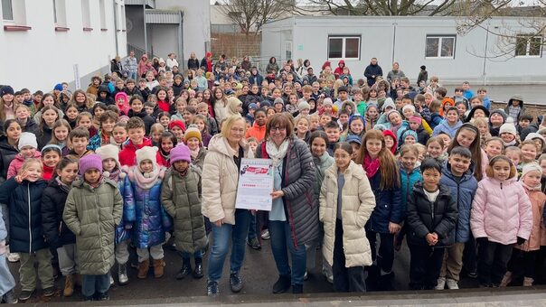 Zwei Frauen stehen vor einer Traube von Kindern auf dem Schulhof einer Schule und halten gemeinsam einen Bilderrahmen mit einem Zertifikat in den Händen.