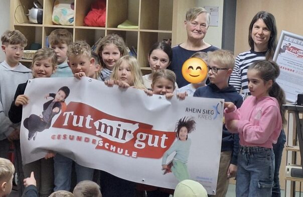 Eine Gruppe von Kindern hält ein weißes Banner mit rote Turmirgut-gesunde Schule Aufschrift in der Hand. Dahinter stehen zwei Frauen.