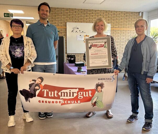 Vier Erwachsene halten ein Banner mit der Aufschrift Tutmirgut gesunde Schule in den Händen.