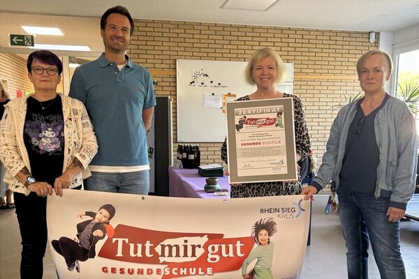 Vier Erwachsene halten ein Banner mit der Aufschrift Tutmirgut gesunde Schule in den Händen.