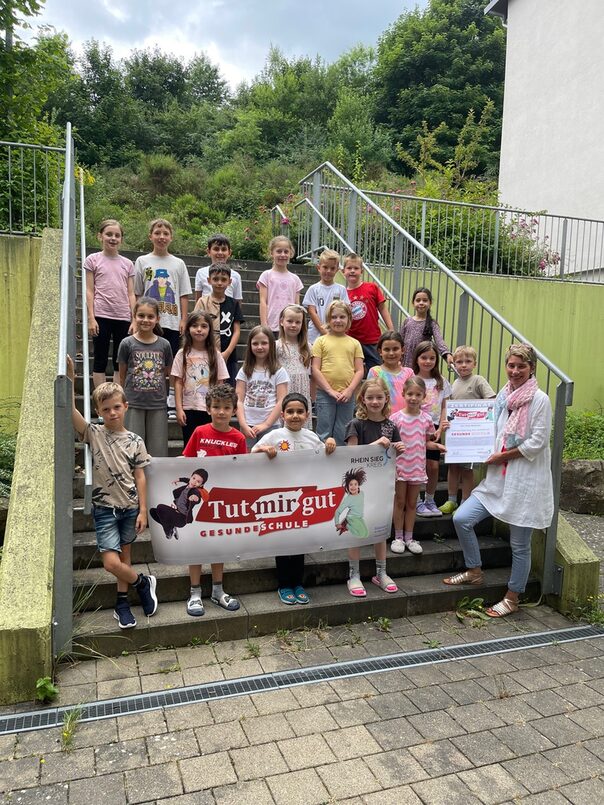 Eine Gruppe von Kindern steht auf einer treppe und hält ein großes Banner mit der Aufschrift Tutmirgut gesunde Schule vor sich. Am Rand steht eine Frau.