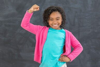 Adorable African American elementary schoolgirl smiles while flexing her biceps. She is standing in front of a chalkboard in a classroom.